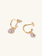 A pair of 15mm diameter gold hoop earrings each with an 8mm purple Bazilian Amethyst gemstone drop and butterfly fastening. Set on a cream background with gentle shadowing.
