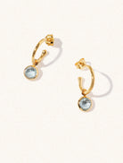 A pair of 15mm diameter gold hoop earrings each with an 8mm blue topaz gemstone drop and butterfly fastening. Set on a cream background with gentle shadowing.
