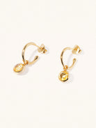 A pair of 15mm diameter gold hoop earrings each with an 8mm citrine gemstone drop and butterfly fastening. Set on a cream background with gentle shadowing.