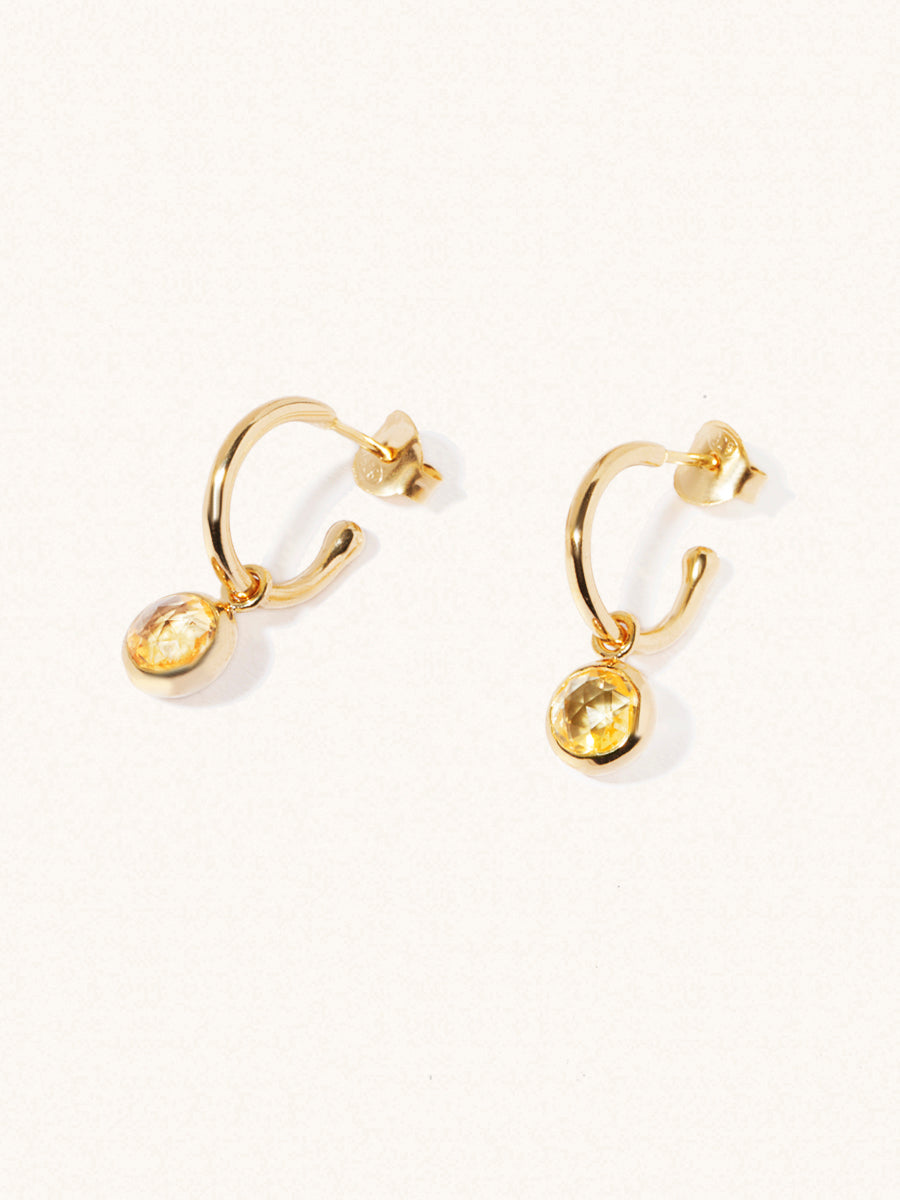 A pair of 15mm diameter gold hoop earrings each with an 8mm citrine gemstone drop and butterfly fastening. Set on a cream background with gentle shadowing.