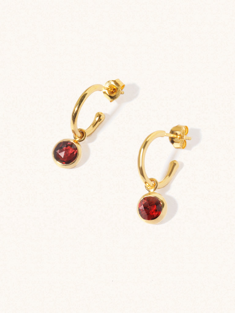 A pair of 15mm diameter gold hoop earrings each with an 8mm deep red Garnet drop gemstone an butterfly fastening. Set on a cream background with gentle shadowing.