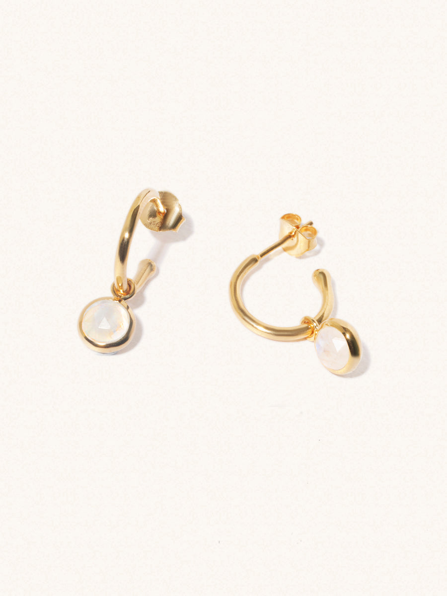 A pair of 15mm diameter gold hoop earrings each with an 8mm moonstone gemstone drop and butterfly fastening. Set on a cream background with gentle shadowing.