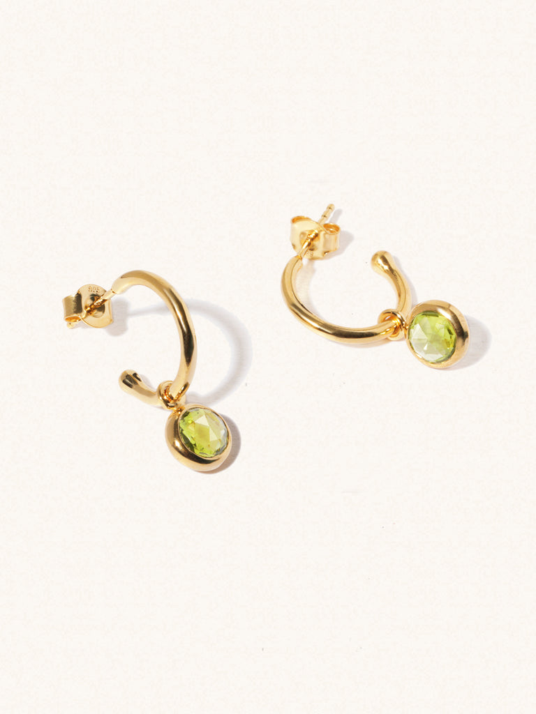 A pair of 15mm diameter gold hoop earrings each with an 8mm green Peridot gemstone drop and butterfly fastening. Set on a cream background with gentle shadowing.