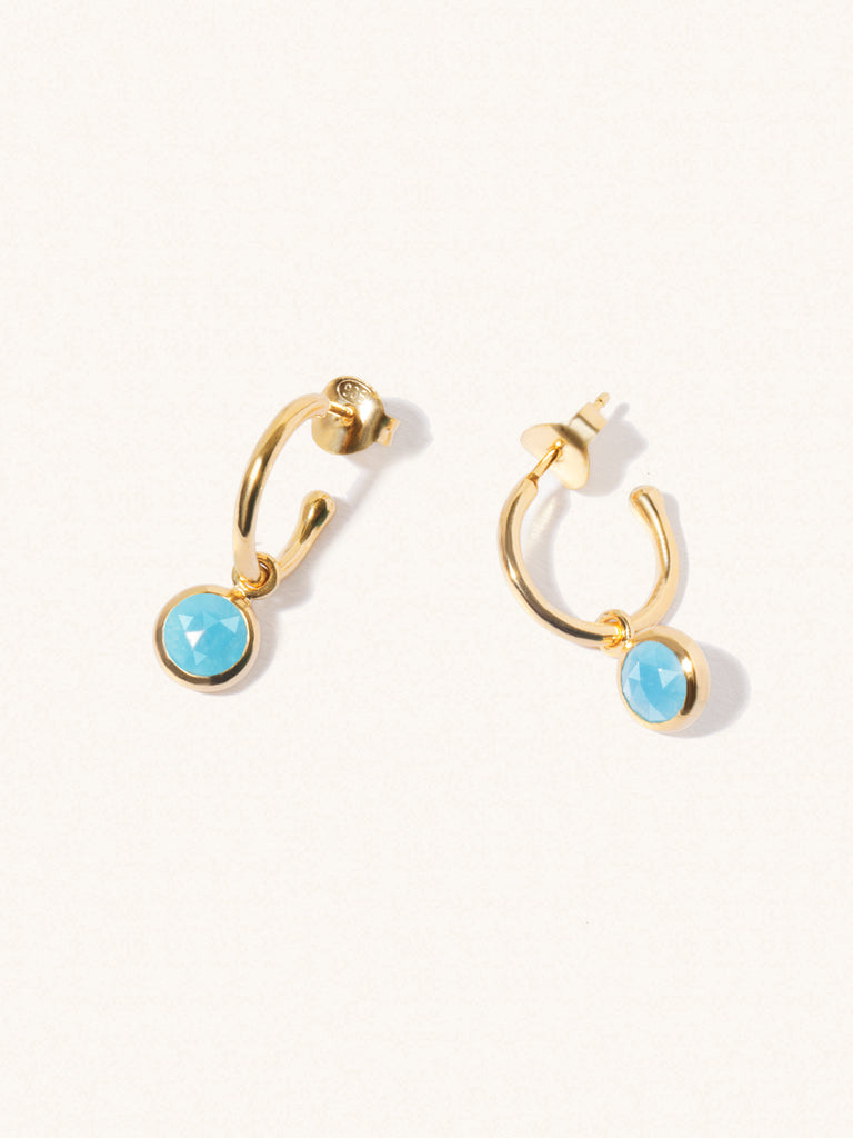 A pair of 15mm diameter gold hoop earrings each with an 8mm turquoise gemstone drop and butterfly fastening. Set on a cream background with gentle shadowing.