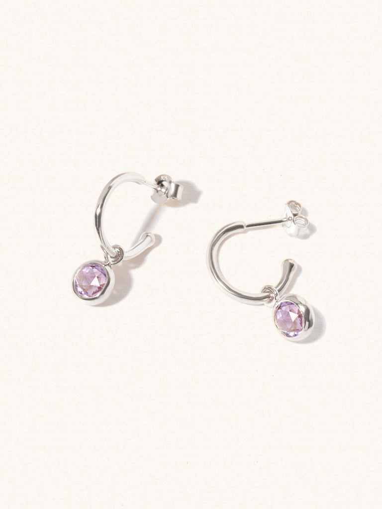 A pair of 15mm diameter gold hoop earrings each with an 8mm purple Bazilian Amethyst gemstone drop and butterfly fastening. Set on a cream background with gentle shadowing.