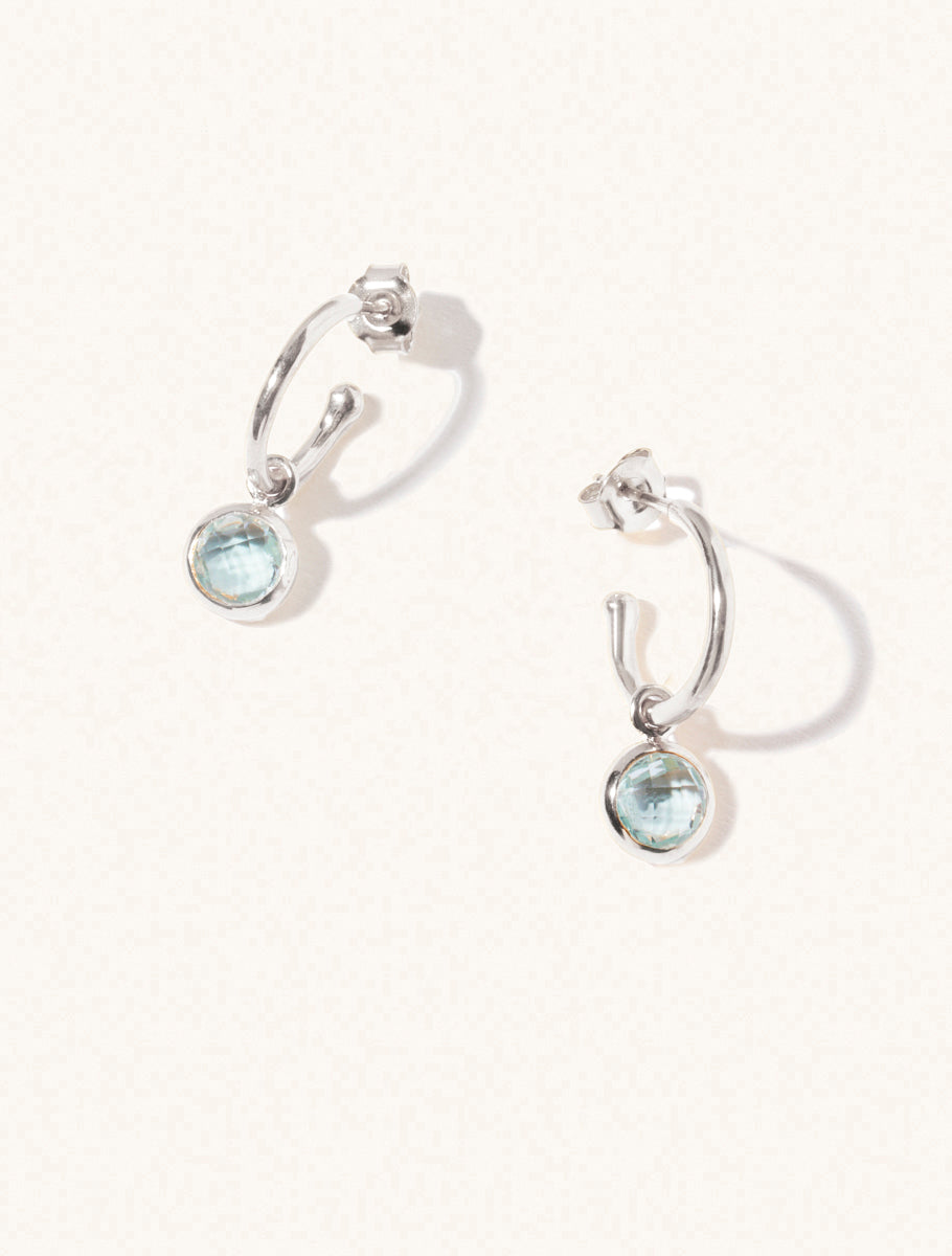 A pair of 15mm diameter sterling silver hoop earrings each with an 8mm blue topaz gemstone drop and butterfly fastening. Set on a cream background with gentle shadowing.
