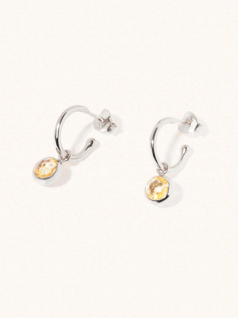 A pair of 15mm diameter sterling silver hoop earrings each with an 8mm citrine gemstone drop and butterfly fastening. Set on a cream background with gentle shadowing.