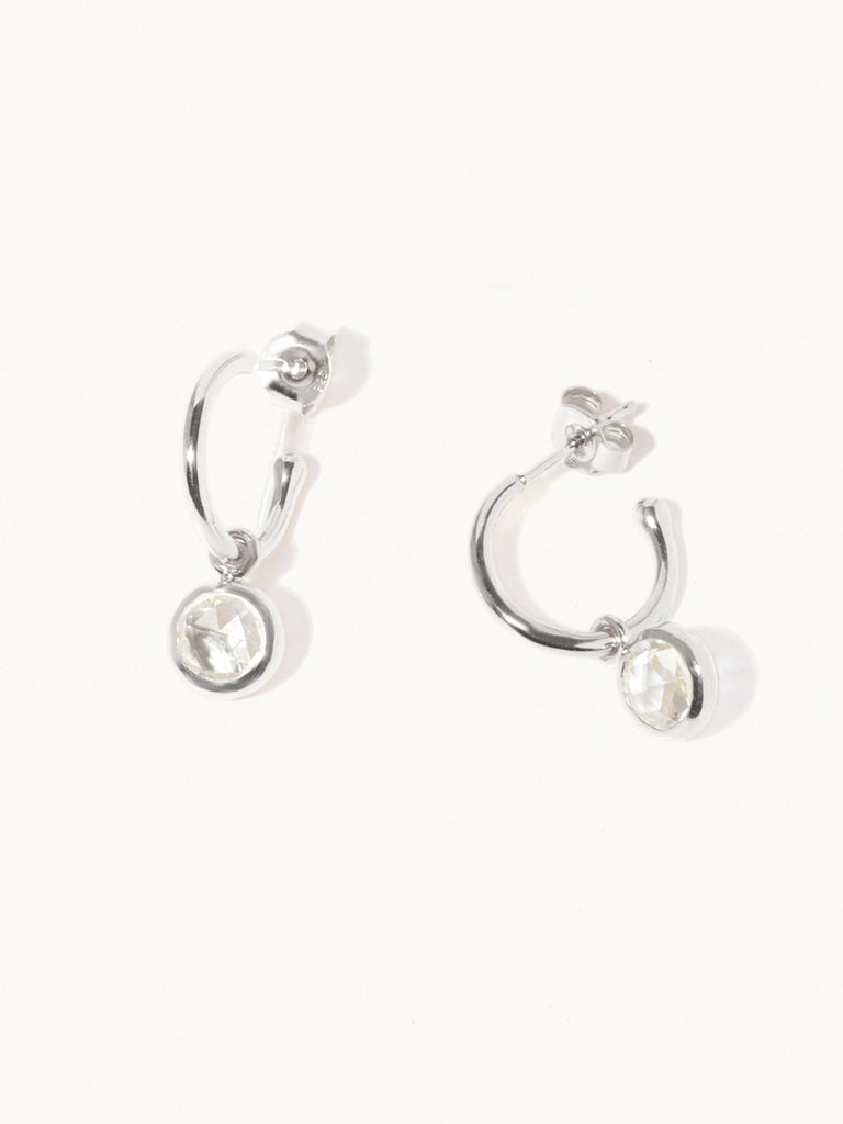 A pair of 15mm diameter sterling silver hoop earrings each with an 8mm deep white crystal gemstone drop and butterfly fastening. Set on a cream background with gentle shadowing.