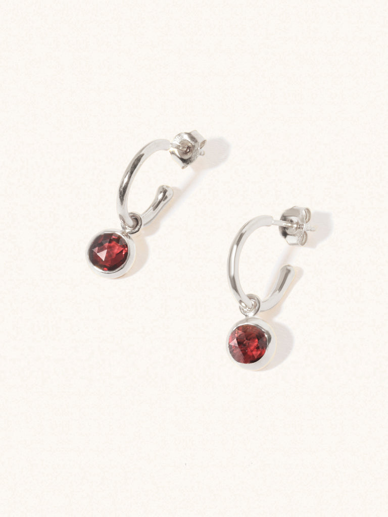 A pair of 15mm diameter sterling silver hoop earrings with an 8mm deep red Garnet drop gemstone. Set on a cream background with gentle shadowing.