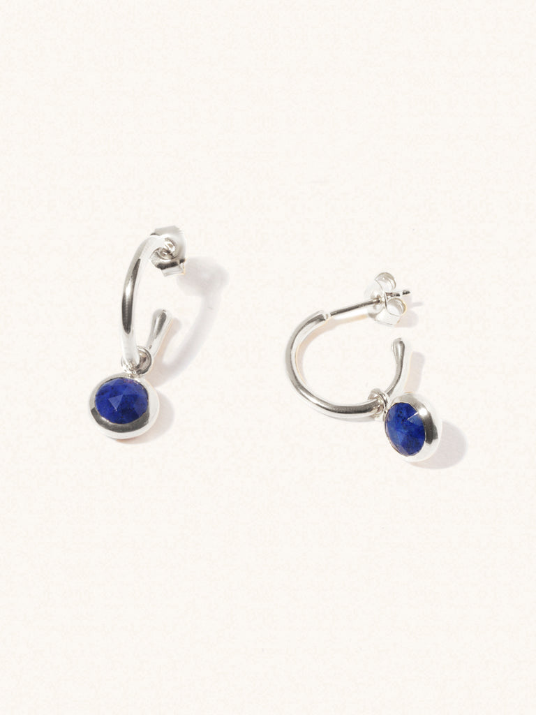 A pair of 15mm diameter sterling silver hoop earrings each with an 8mm lapsi lazuli gemstone drop and butterfly fastening. Set on a cream background with gentle shadowing.