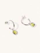 A pair of 15mm diameter sterling silver hoop earrings each with an 8mm green Peridot gemstone drop and butterfly fastening. Set on a cream background with gentle shadowing.