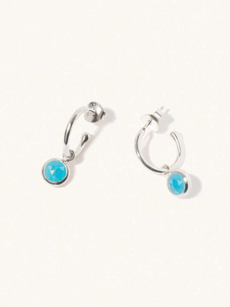 A pair of 15mm diameter sterling silver hoop earrings each with an 8mm turquoise gemstone drop and butterfly fastening. Set on a cream background with gentle shadowing.