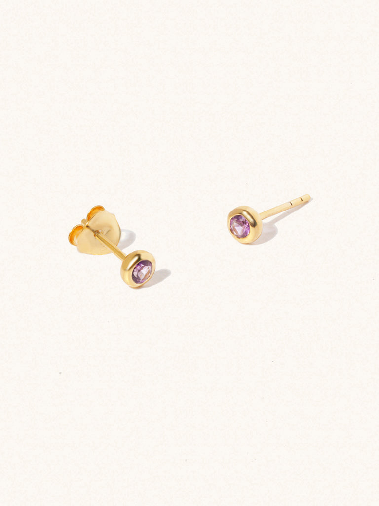 A pair of gold small stud earrings with a 3mm purple Brazilian Amethyst gemstone in a 4mm setting and butterfly fastening. Set on a cream background with gentle shadowing.