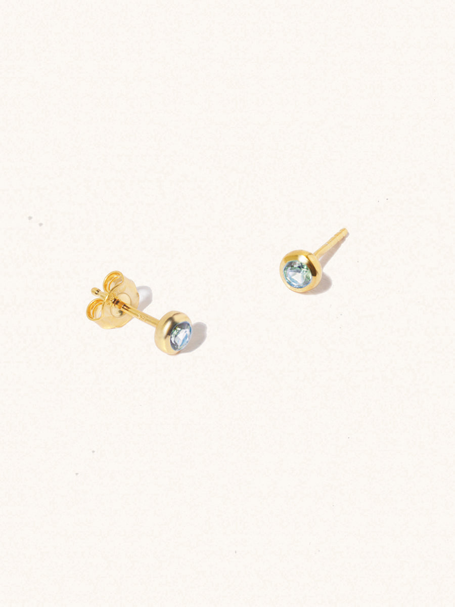 A pair of gold small stud earrings with a 3mm blue topaz gemstone in a 4mm setting and butterfly fastening. Set on a cream background with gentle shadowing.