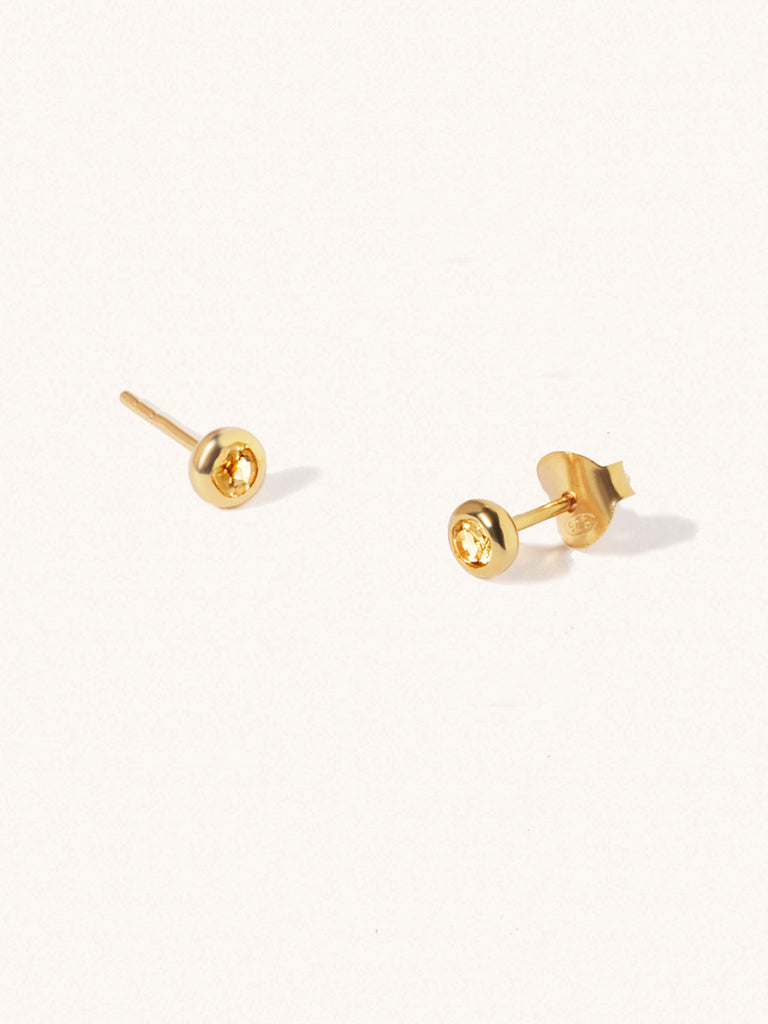 A pair of gold small stud earrings with a 3mm yellow citrine gemstone in a 4mm setting and butterfly fastening. Set on a cream background with gentle shadowing.