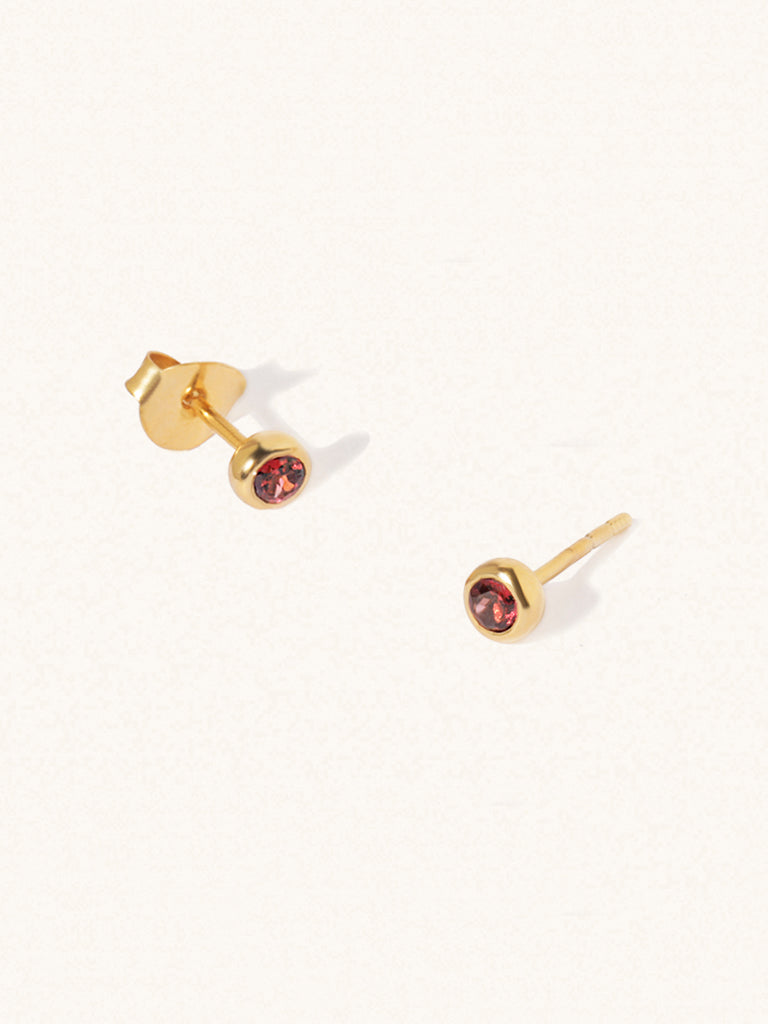 A pair of gold small stud earrings with a 3mm deep red Garnet gemstone in a 4mm setting and butterfly fastening. Set on a cream background with gentle shadowing.