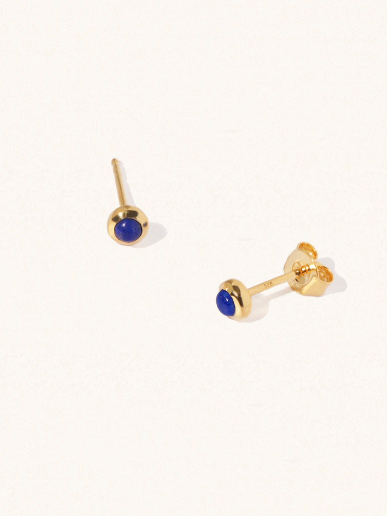 A pair of gold small stud earrings with a 3mm lapis lazuli gemstone in a 4mm setting and butterfly fastening. Set on a cream background with gentle shadowing.