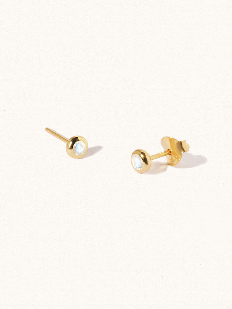 A pair of gold small stud earrings with a 3mm moonstone gemstone in a 4mm setting and butterfly fastening. Set on a cream background with gentle shadowing.