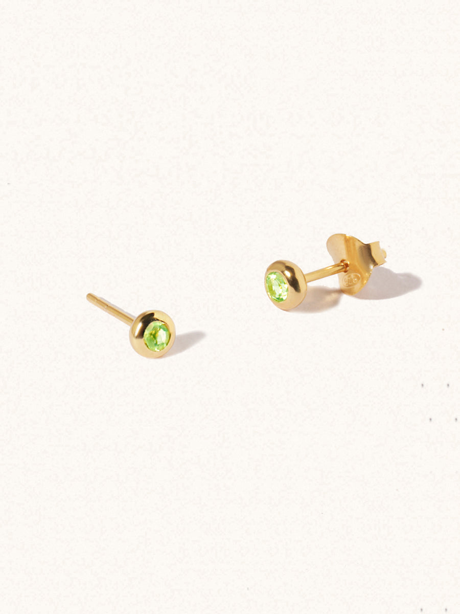 A pair of gold small stud earrings with a 3mm green Peridot gemstone in a 4mm setting and butterfly fastening. Set on a cream background with gentle shadowing.