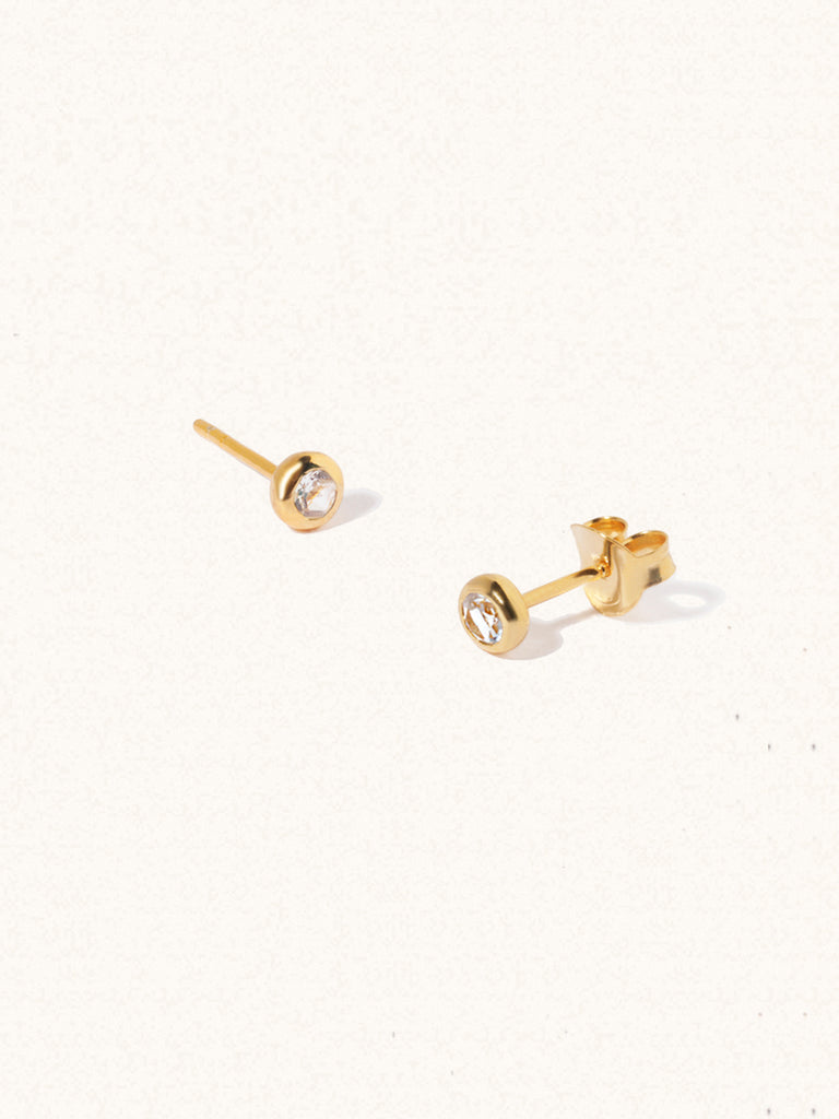 A pair of gold small stud earrings with a 3mm white topaz gemstone in a 4mm setting and butterfly fastening. Set on a cream background with gentle shadowing.