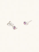 A pair of sterling silver small stud earrings with a 3mm purple Brazilian Amethyst gemstone in a 4mm setting and butterfly fastening. Set on a cream background with gentle shadowing.