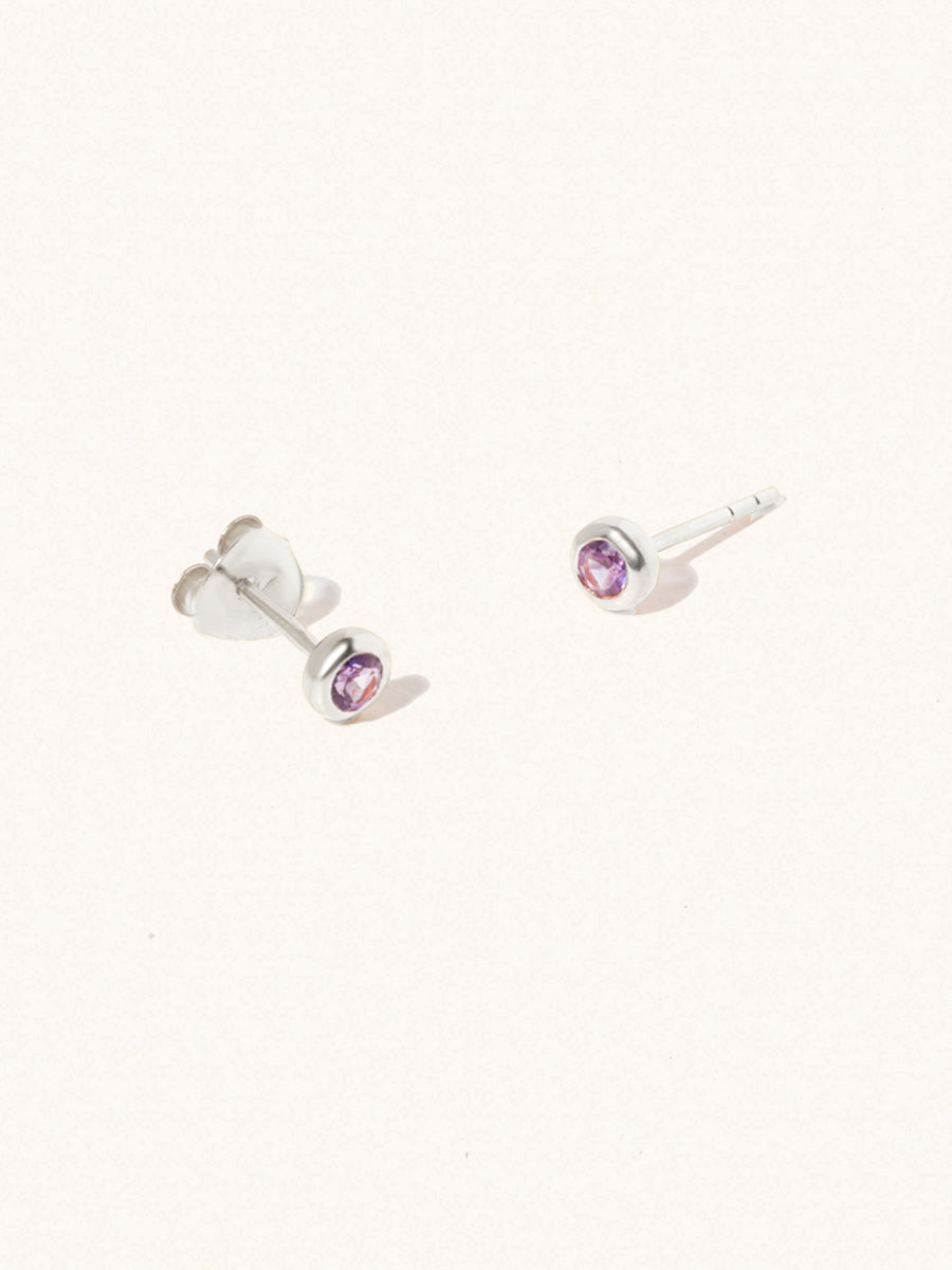 A pair of sterling silver small stud earrings with a 3mm purple Brazilian Amethyst gemstone in a 4mm setting and butterfly fastening. Set on a cream background with gentle shadowing.