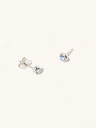 A pair of sterling silver small stud earrings with a 3mm blue topaz gemstone in a 4mm setting and butterfly fastening. Set on a cream background with gentle shadowing.