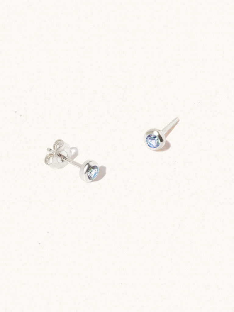 A pair of sterling silver small stud earrings with a 3mm blue topaz gemstone in a 4mm setting and butterfly fastening. Set on a cream background with gentle shadowing.