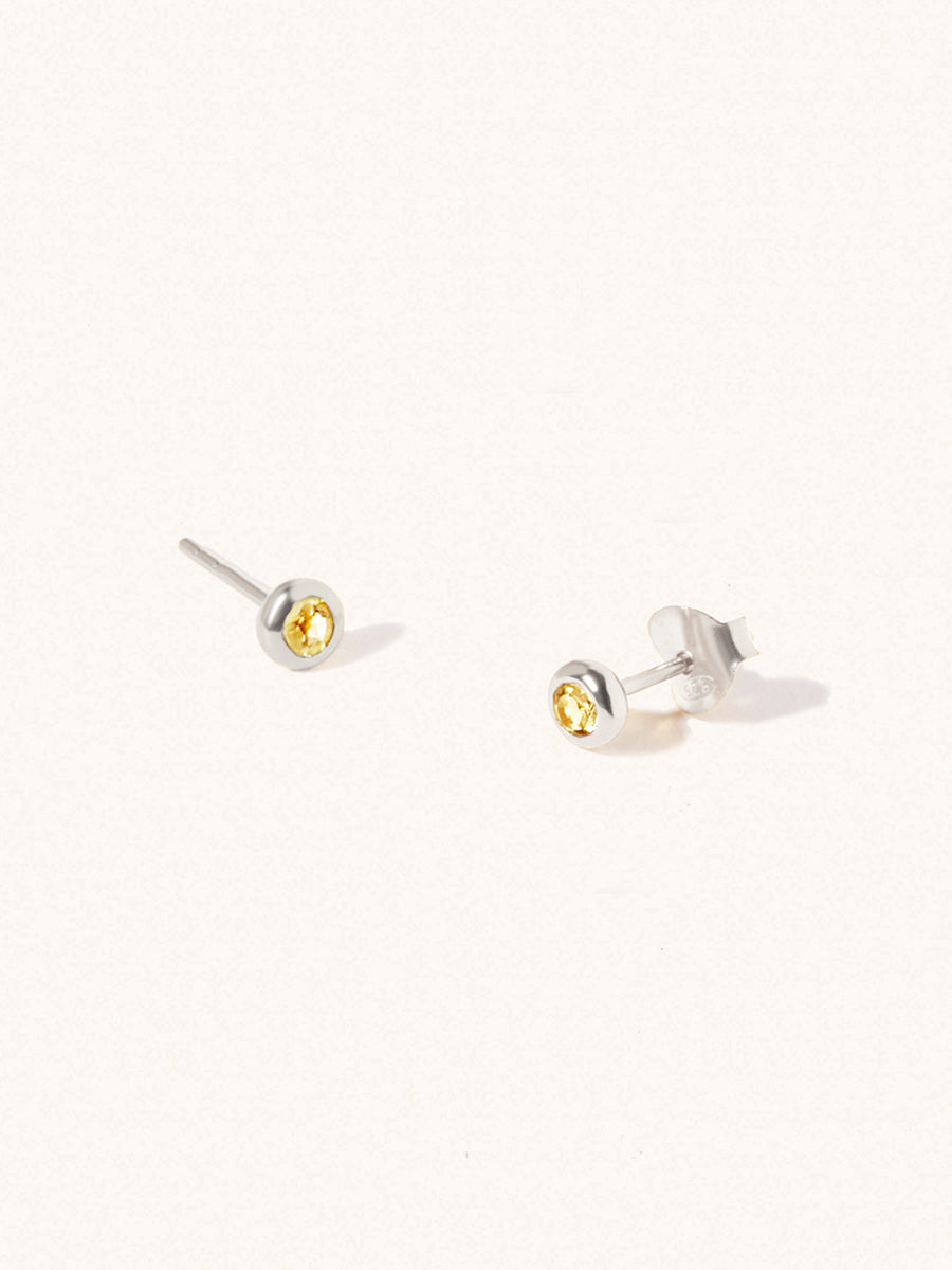 A pair of sterling silver small stud earrings with a 3mm yellow citrine gemstone in a 4mm setting and butterfly fastening. Set on a cream background with gentle shadowing.