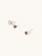 A pair of sterling silver small stud earrings with a 3mm deep red Garnet gemstone in a 4mm setting and butterfly fastening. Set on a cream background with gentle shadowing.