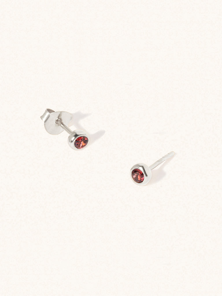 A pair of sterling silver small stud earrings with a 3mm deep red Garnet gemstone in a 4mm setting and butterfly fastening. Set on a cream background with gentle shadowing.