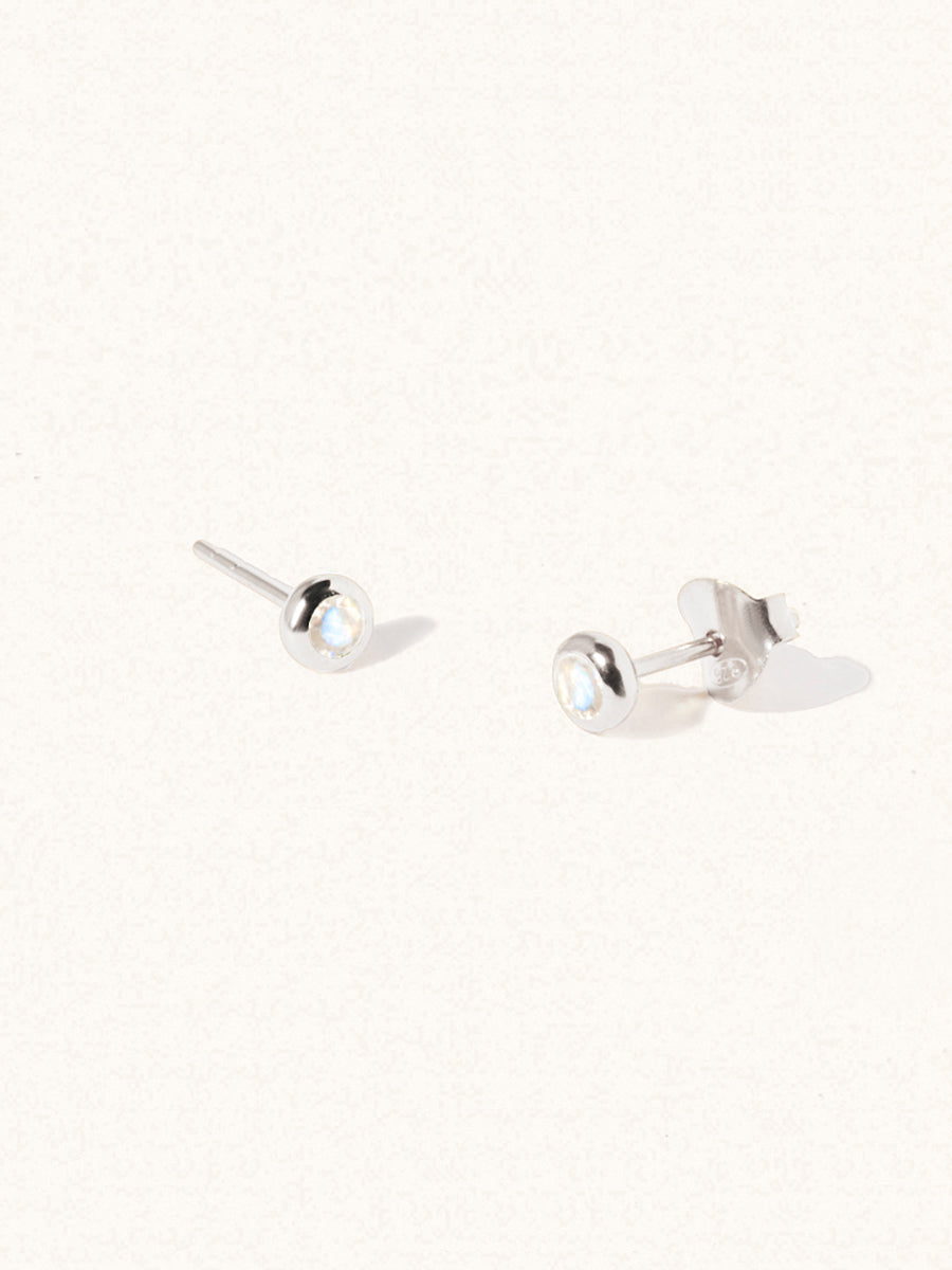 A pair of sterling silver small stud earrings with a 3mm moonstone gemstone in a 4mm setting and butterfly fastening. Set on a cream background with gentle shadowing.