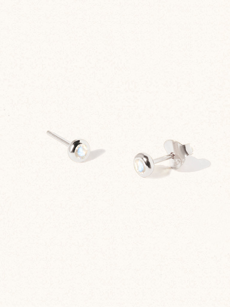 A pair of sterling silver small stud earrings with a 3mm moonstone gemstone in a 4mm setting and butterfly fastening. Set on a cream background with gentle shadowing.