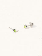 A pair of sterling silver small stud earrings with a 3mm green Peridot gemstone in a 4mm setting and butterfly fastening. Set on a cream background with gentle shadowing.