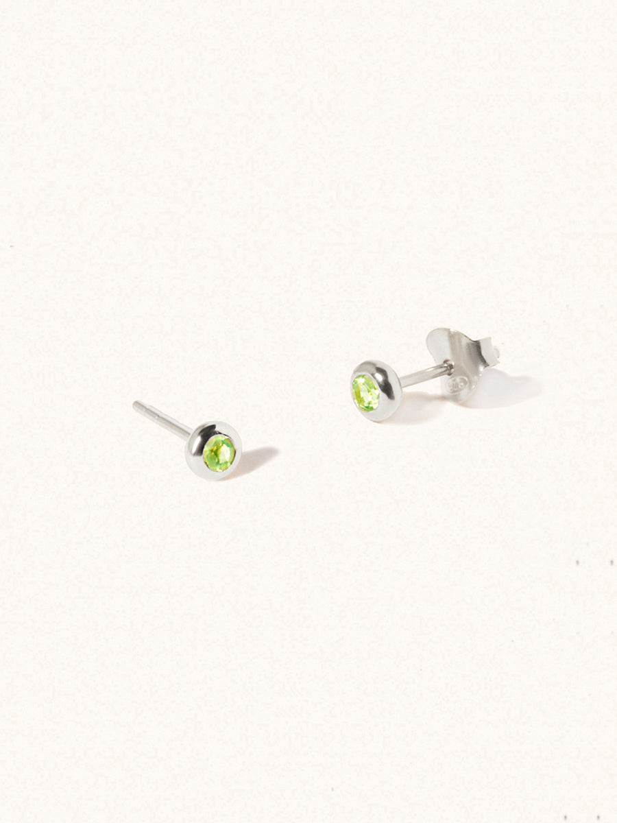 A pair of sterling silver small stud earrings with a 3mm green Peridot gemstone in a 4mm setting and butterfly fastening. Set on a cream background with gentle shadowing.