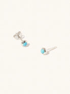 A pair of sterling silver small stud earrings with a 3mm turquoise gemstone in a 4mm setting and butterfly fastening. Set on a cream background with gentle shadowing.