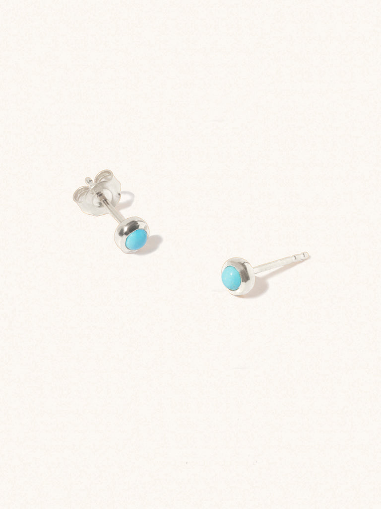 A pair of sterling silver small stud earrings with a 3mm turquoise gemstone in a 4mm setting and butterfly fastening. Set on a cream background with gentle shadowing.