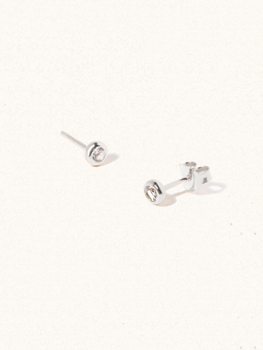 A pair of sterling silver small stud earrings with a 3mm white topaz gemstone in a 4mm setting and butterfly fastening. Set on a cream background with gentle shadowing.