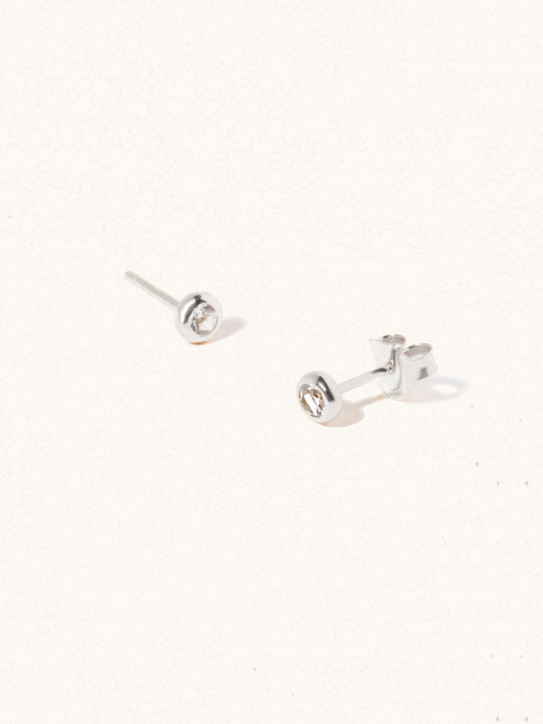 A pair of sterling silver small stud earrings with a 3mm white topaz gemstone in a 4mm setting and butterfly fastening. Set on a cream background with gentle shadowing.