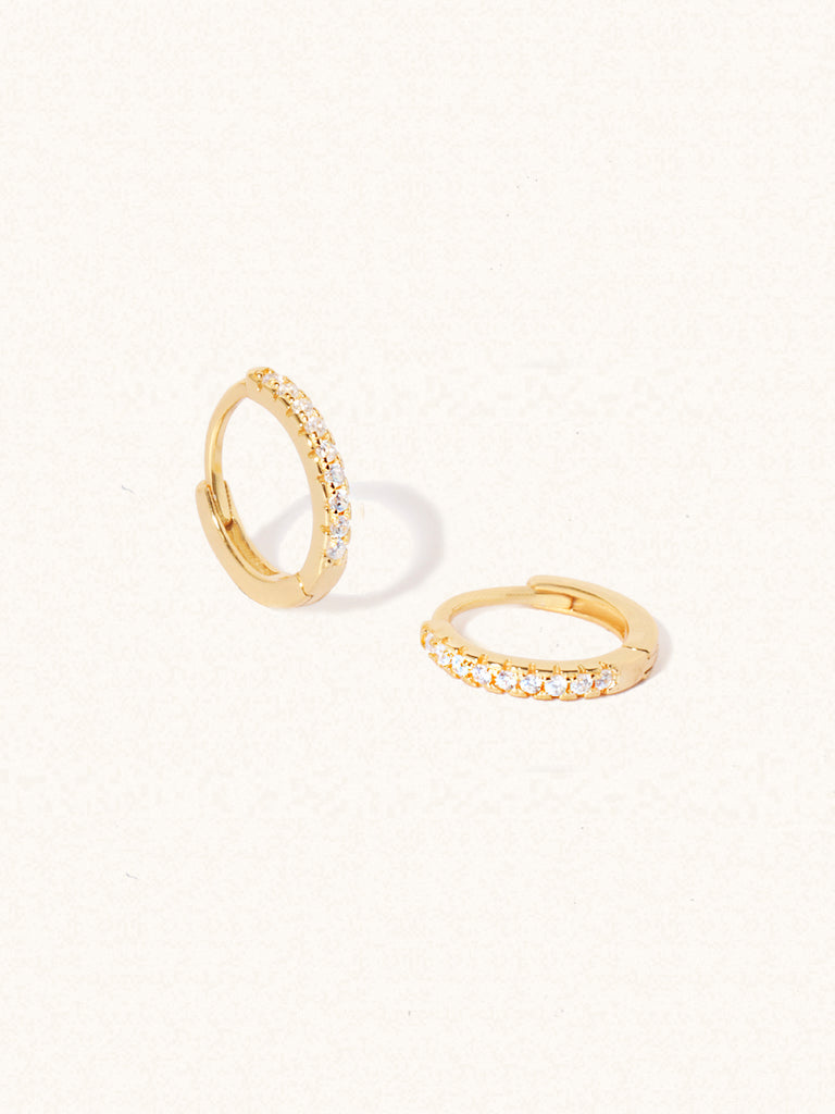 A pair of gold plated huggies with 10 white topaz gemstones per huggie. Set on a cream background with elongated shadows