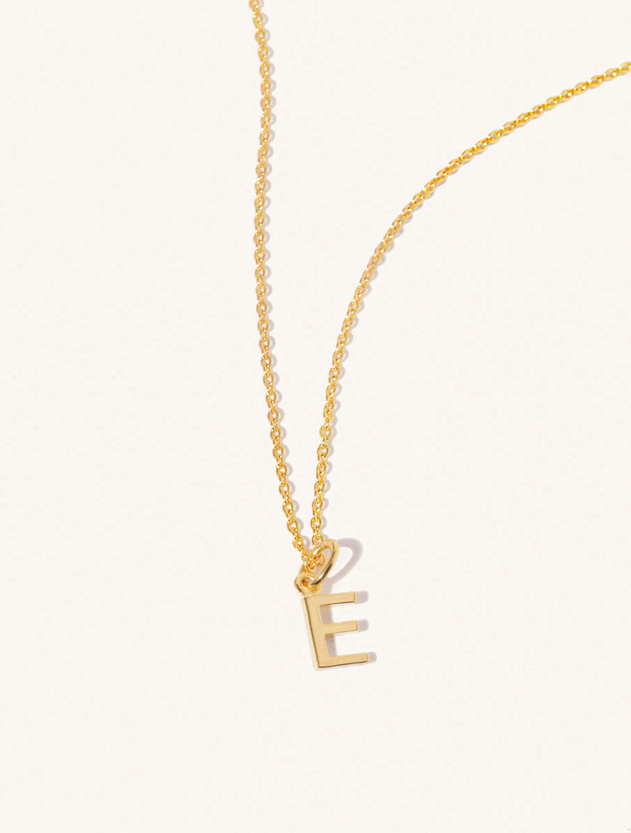 A gold chain necklace with the caplital letter E approximately 8mm x 6mm. Set on a cream background with gentle shadowing.