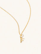 A gold chain necklace with the caplital letter F approximately 8mm x 6mm. Set on a cream background with gentle shadowing.