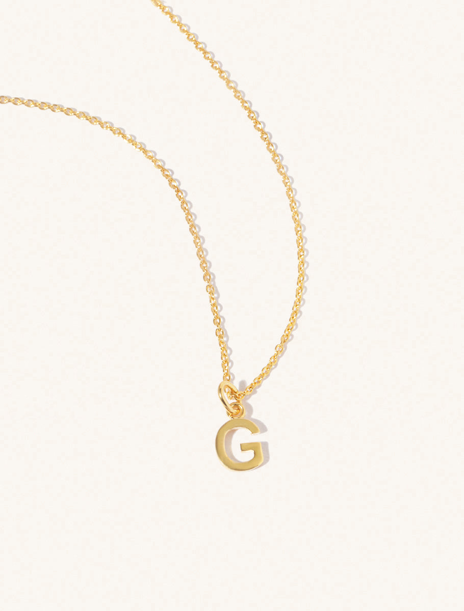 A gold chain necklace with the caplital letter G approximately 8mm x 6mm. Set on a cream background with gentle shadowing.