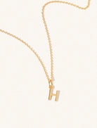 A gold chain necklace with the caplital letter H approximately 8mm x 6mm. Set on a cream background with gentle shadowing.