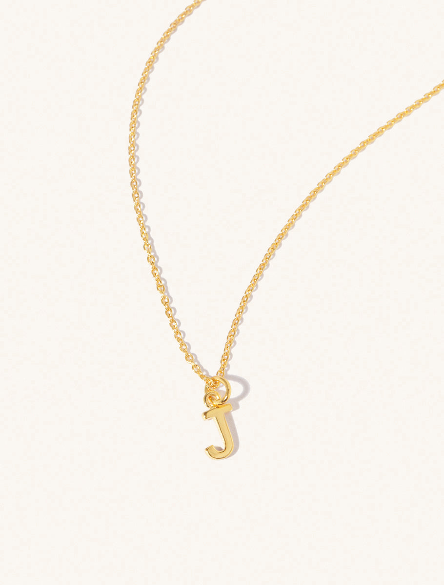 A gold chain necklace with the caplital letter J approximately 8mm x 6mm. Set on a cream background with gentle shadowing.