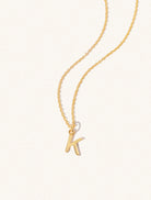 A gold chain necklace with the caplital letter K approximately 8mm x 6mm. Set on a cream background with gentle shadowing.