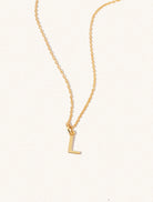 A gold chain necklace with the caplital letter L approximately 8mm x 6mm. Set on a cream background with gentle shadowing.