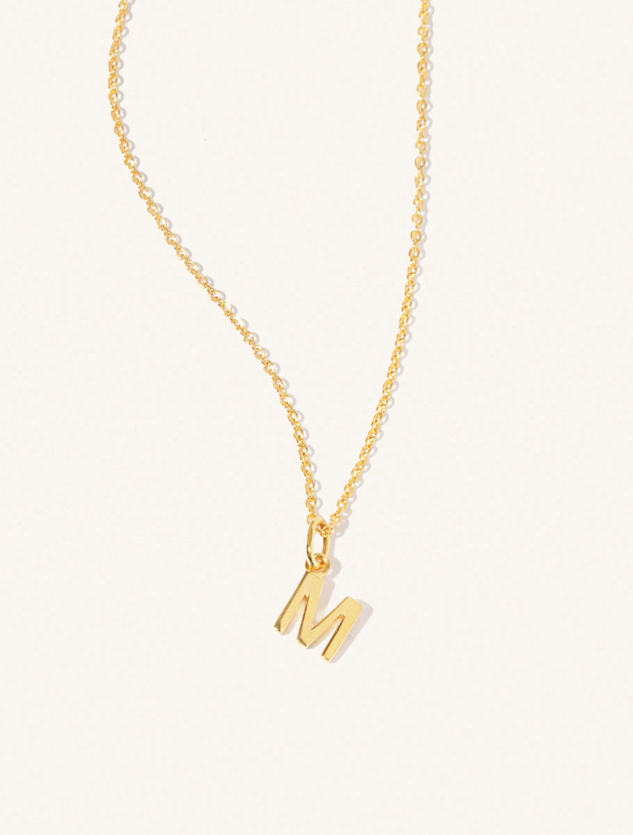 A gold chain necklace with the caplital letter M approximately 8mm x 6mm. Set on a cream background with gentle shadowing.