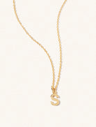 A gold chain necklace with the caplital letter S approximately 8mm x 6mm. Set on a cream background with gentle shadowing.