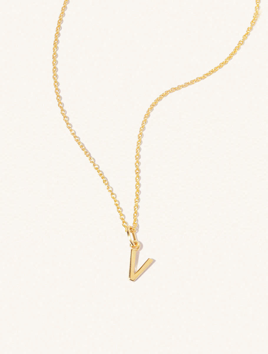 A gold chain necklace with the caplital letter V approximately 8mm x 6mm. Set on a cream background with gentle shadowing.